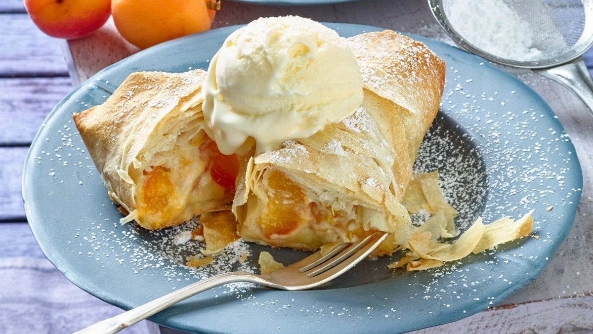 Eine angeschnittene Mini-Aprikosen-Strudelrolle liegt auf einem blauen Teller auf einem hölzernen Untergrund. Neben der Rolle liegt eine silberne Gabel. Die Strudelrolle ist garniert mit Puderzucker und einer Kugel Vanilleeis. Im Hintergrund liegt ein Sieb mit Puderzucker und zwei Aprikosen.