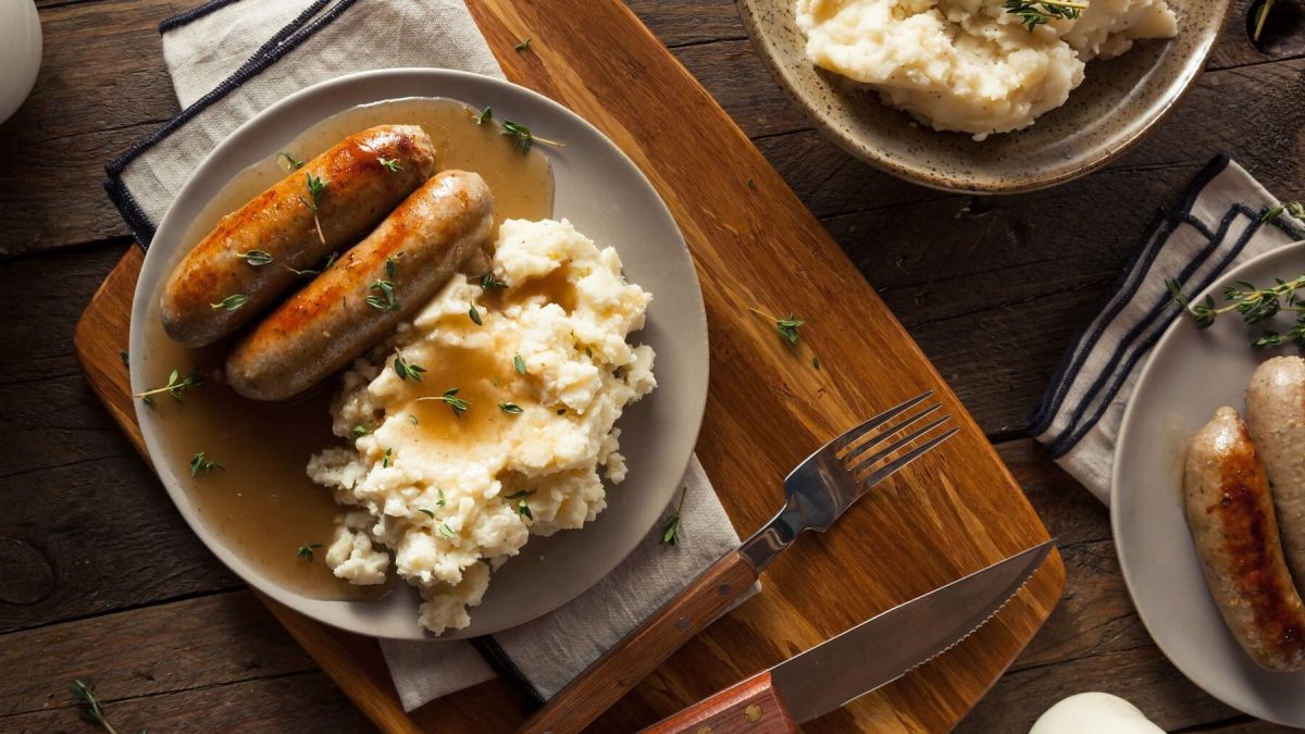 Bangers and Mash von oben auf einem hellrauen Tellern neben Besteck auf einem dunklen Holztisch.
