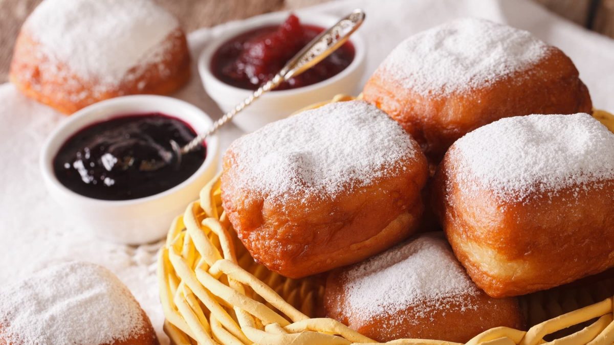 Gebackene Beignets mit Puderzucker in einem hellen Korb vor Schüsseln mit Marmelade auf einem Tisch mit weißer Tischdecke.