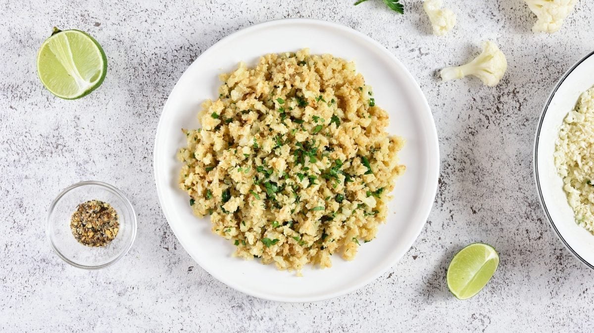 Ein Teller mit gebratenem und mit Kräutern gewürztem Blumenkohlreis neben Limetten Scheiben und Blumenkohlröschen, von oben vor einem grauen Hintergrund.