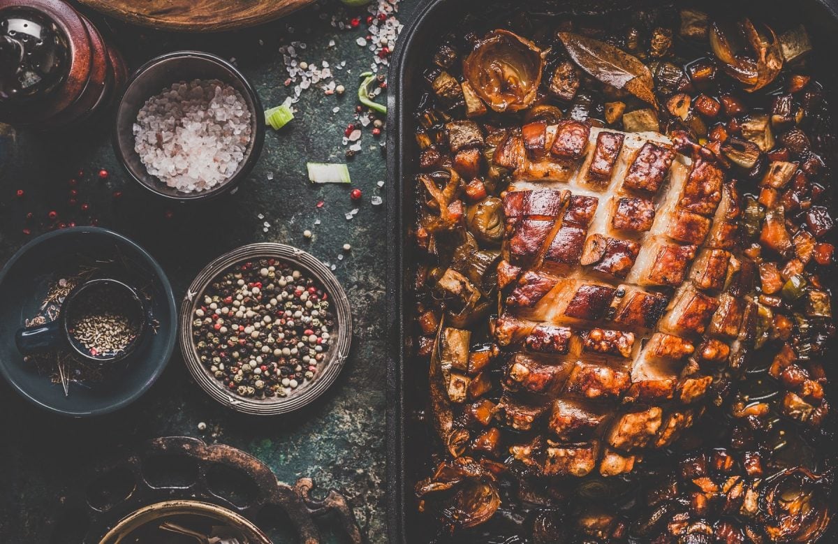 Ein Braten mit ziselierter Kruste liegt auf einer Schieferplatte mit allerlei Gewürzen drumherum