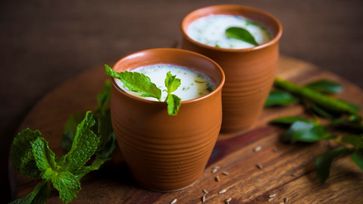 Zwei Ton-Tassen gefüllt mit dem schaumigen Buttermilch-Lassi mit Minze garniert, auf einem Holzbrett von der Seite.