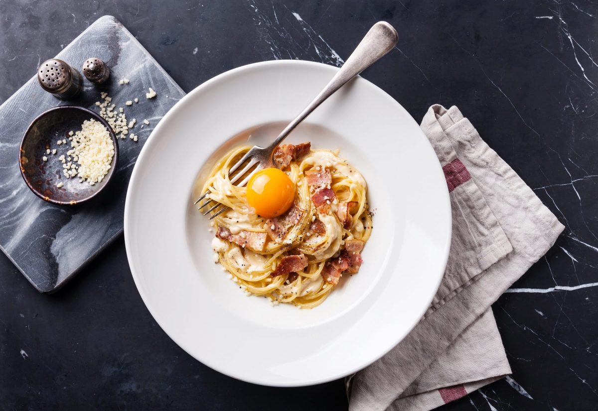 Ein weißer Teller mit Spgehtti Carbonara, Stoffserviette und einer Schale mit geriebenem Käse