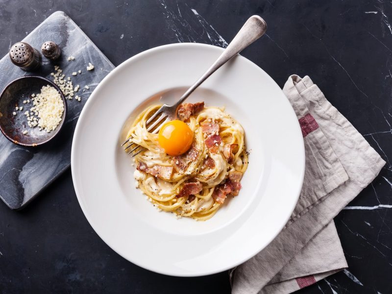 Ein weißer Teller mit Spgehtti Carbonara, Stoffserviette und einer Schale mit geriebenem Käse