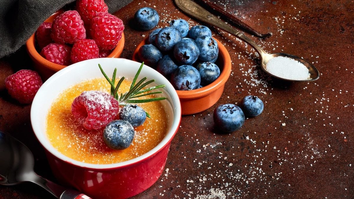 Crème Brûlée mit Kardamom und Banane in einer roten Form mit frischen Früchten auf einem dunklen Tisch der mit Zucker bestreut ist.