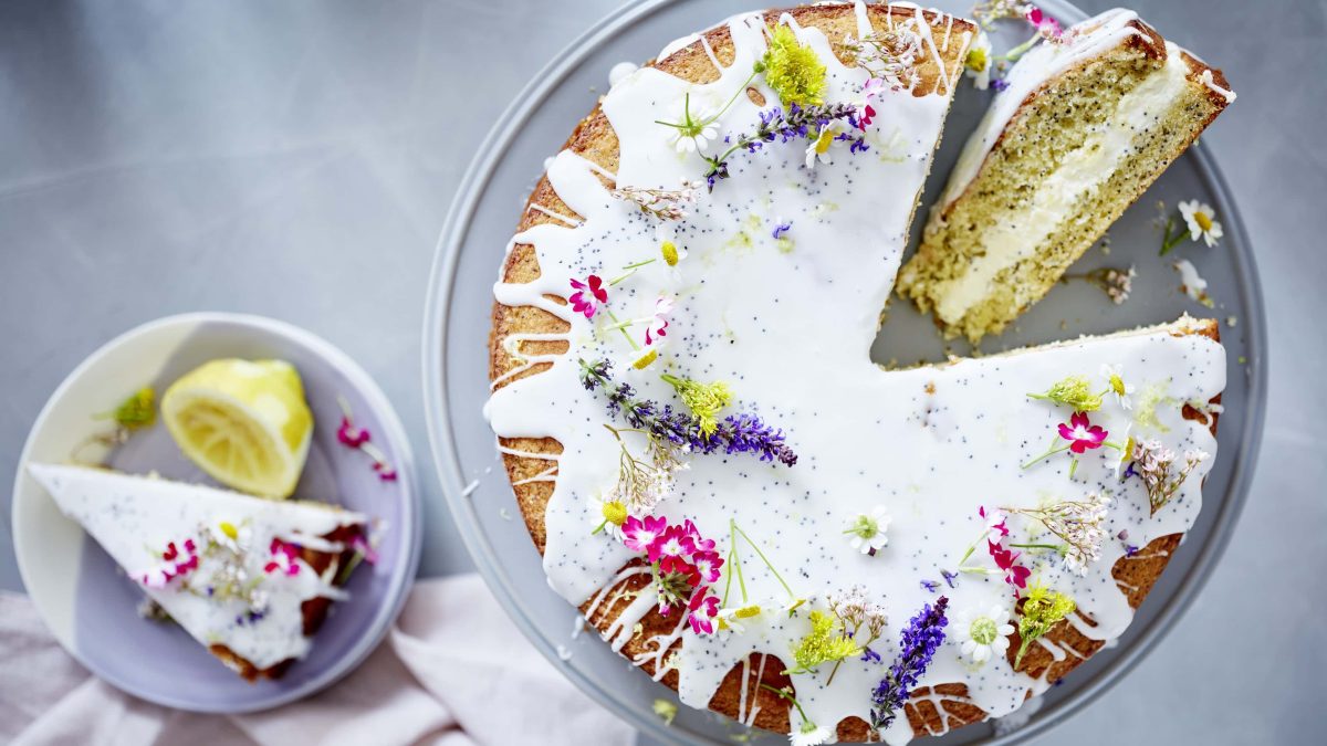 Drizzle-Kuchen mit Zitrone und Mohn verziert mit Blumen. Das Ganze auf einem Tortenhalter und daneben ein Stück auf einem Teller mit einer Zitrone.