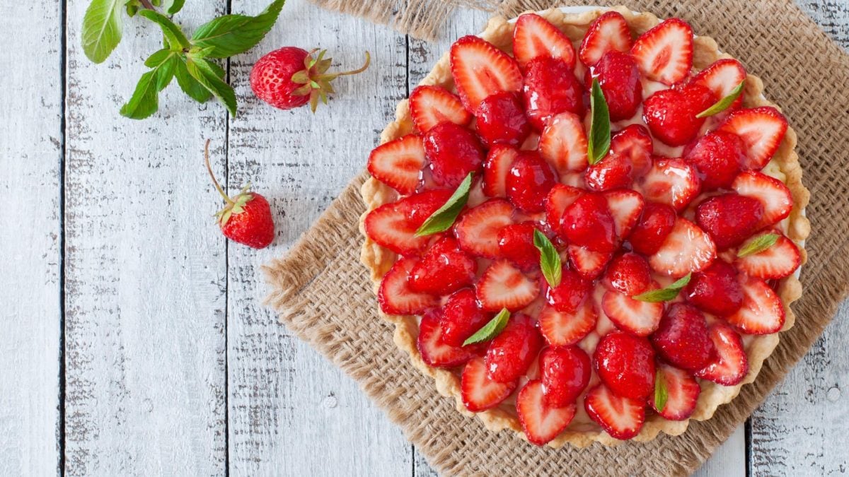Erdbeerkuchen auf einem geflochtenem Tuch. Daneben Minze und Erdbeeren. Das Ganze auf einem hölzernen Untergrund.