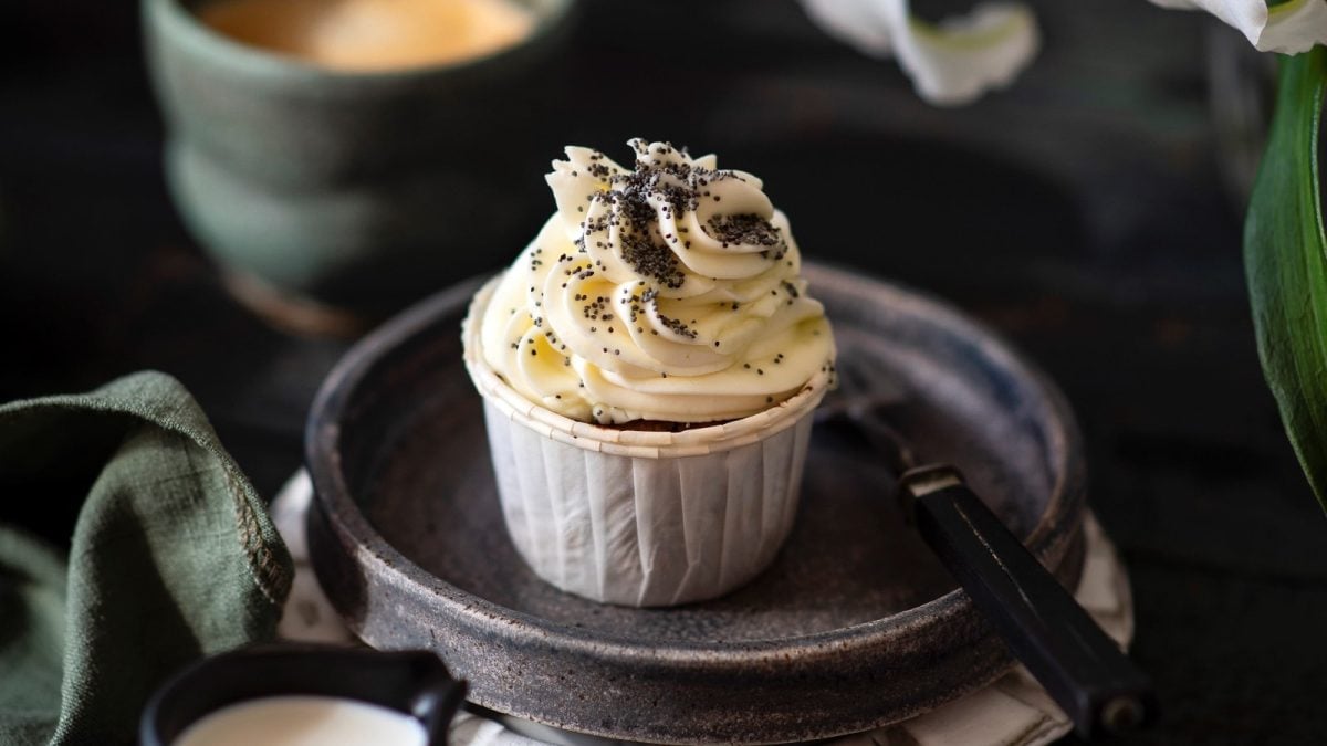 Ein Espresso-Cupcake mit Mohn auf einem dunkelgrauen Teller mit einer Gabel im Vordergrund, sowie eine Tasse Kaffee im Hintergrund.