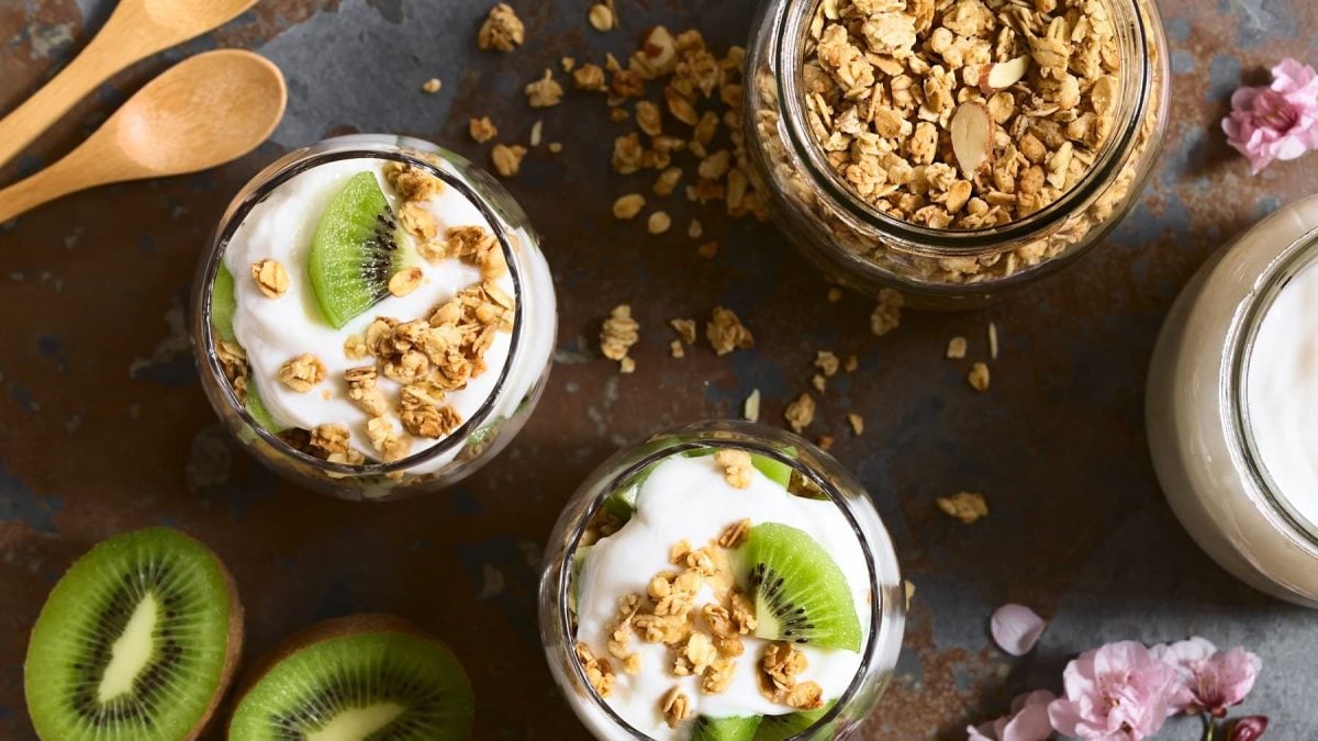Frühstücks-Skyr mit Granola und Kiwi von Oben in Gläsern neben frischer Kiwi und Blüten auf einem grauen Steintisch