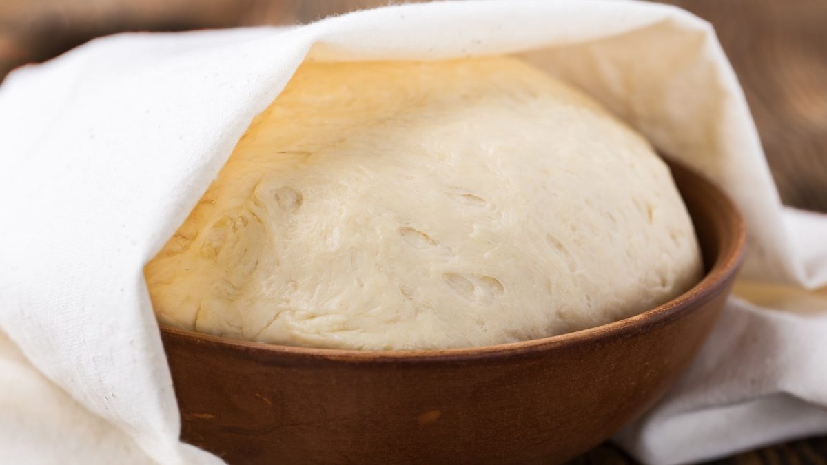 Eine Kugel Hefeteig wird gehen gelassen: Der Teig befindet sich in einer Keramik-Schüssel und ist zur Hälfte mit einem Tuch bedeckt.