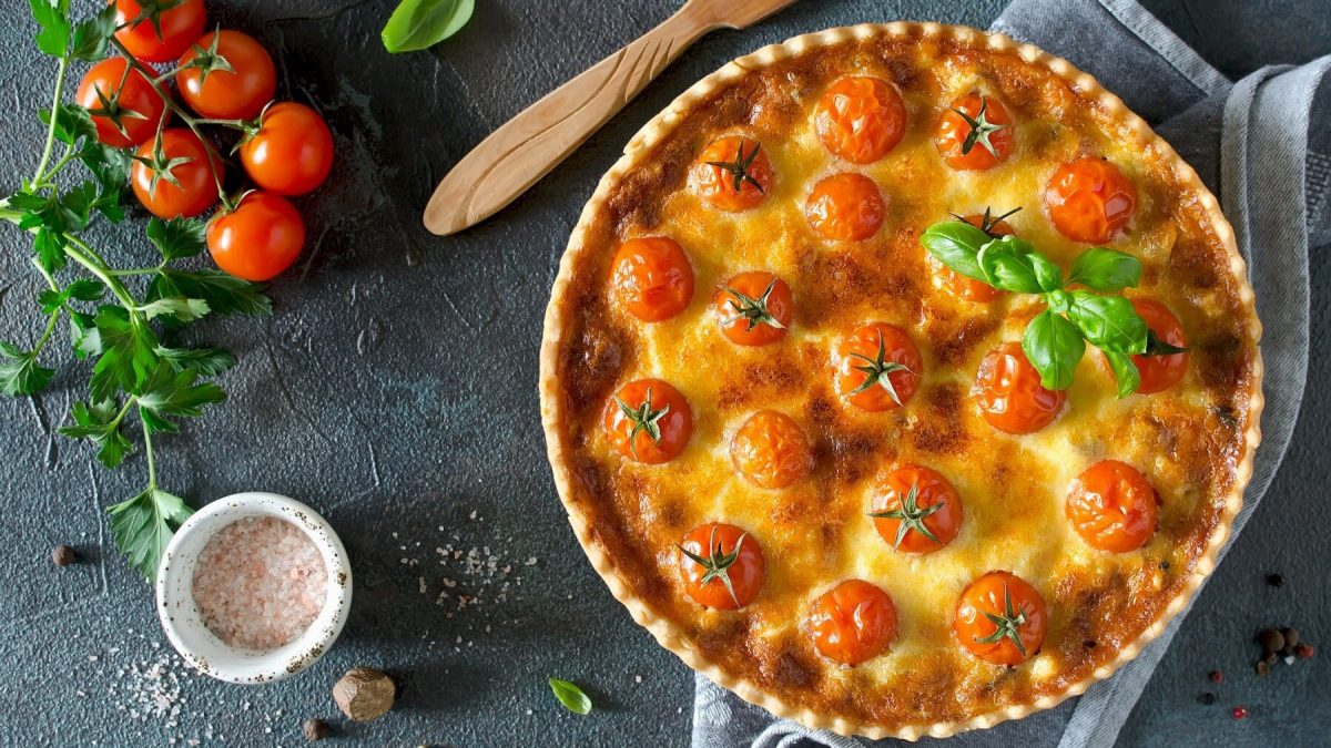 Eine runde Kirschtomaten-Quiche steht auf einem grauen Handtuch auf einem grauen Untergrund. Neben der Quiche liegt ein brauner Holzlöffel. Auf dem Untergrund sind Basilikum, Kirschtomaten und Salzkörner verteilt. Etwas Petersilie liegt neben den Tomaten. Die Quiche ist mit einigen Basilikumblättern angerichtet.