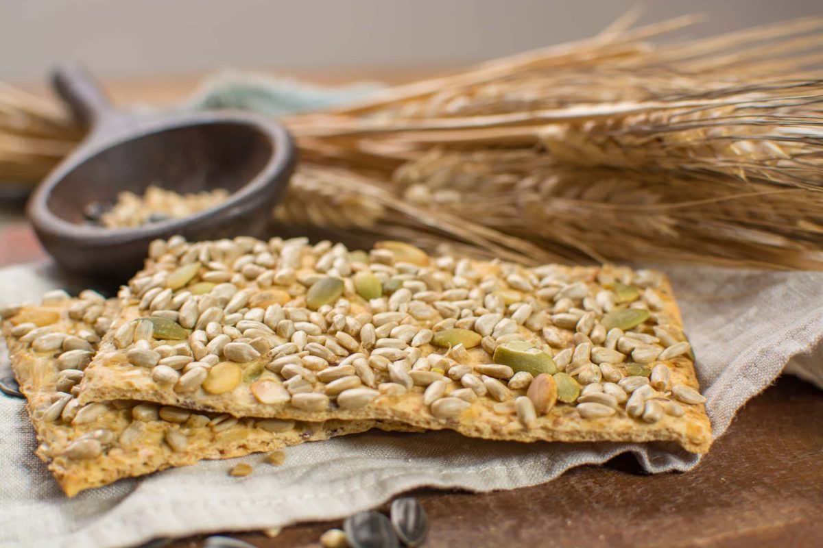 Knäckebrot im Öko-Test: zwei Scheiben Knäckebrot mit Körnern, im Hintergrund Getreide und ein Mörser.