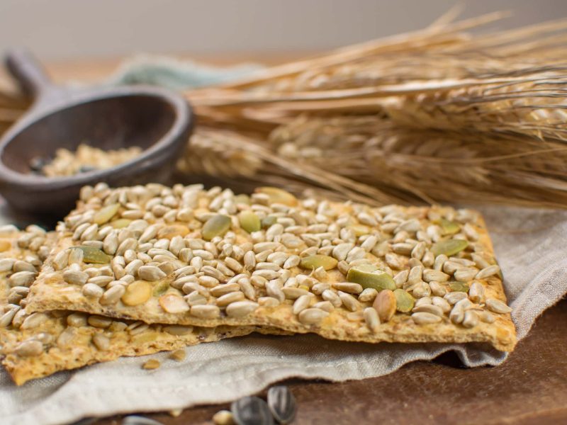 Knäckebrot im Öko-Test: zwei Scheiben Knäckebrot mit Körnern, im Hintergrund Getreide und ein Mörser.