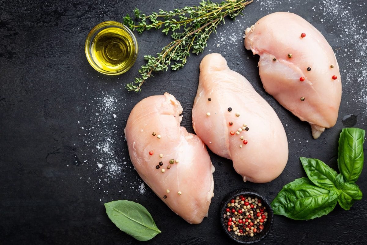 Rohes Hähnchenfleisch für eine Farce von oben auf einem schwarzen Tisch mit frischen Pfefferkörnern, Basilikum, Salz, Thymian und einem Glas Öl.