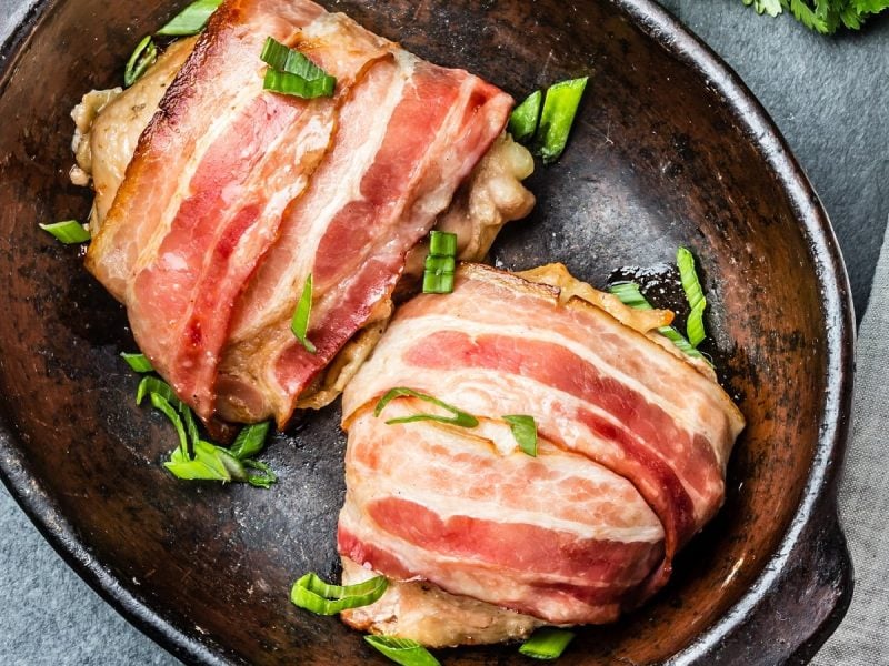 Fleisch bardieren: Zwei bardierte Hühnerfilets in Speck eingewickelt und mit Kräutern besträut in einer Schale, von oben.