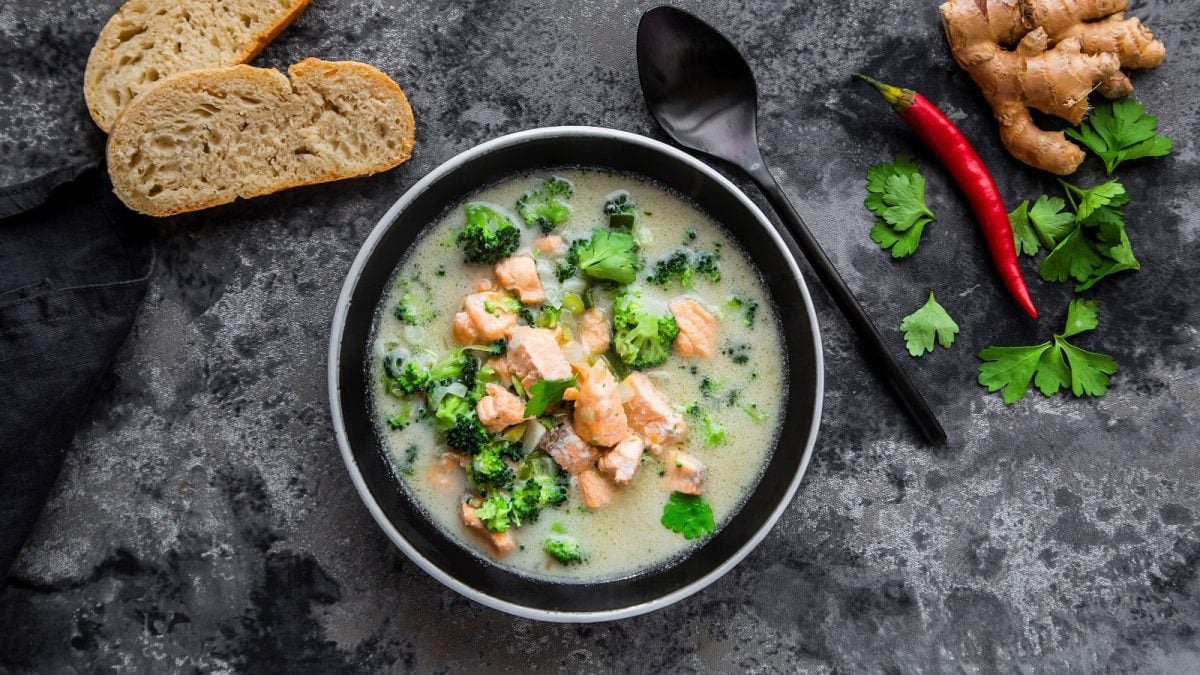 Eine Schüssel mit der Kokos-lachssuppe mit Brokkoli von oben vor grauem Hintergrund neben einer Chilischote, Koriander, Ingwer und zwei Baguette Stücken.