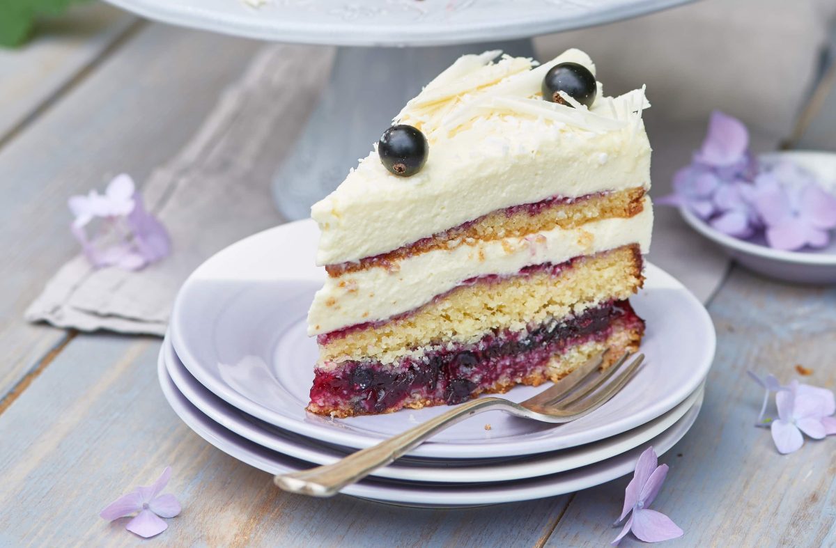 Stück einer Kokostorte mit Johannisbeeren auf weißen Tellern mit einer Gabel auf einem hellen Tisch mit violetten Blüten.
