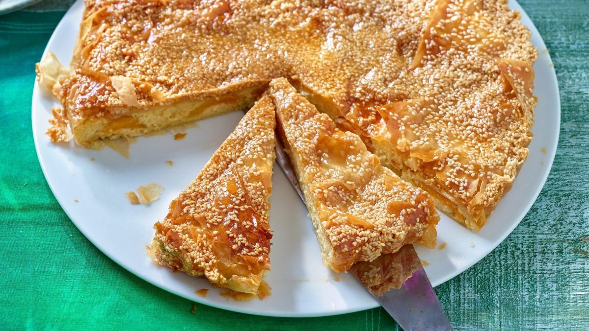 Eine Mandel-Orangen-Tarte steht auf einem weißen Teller. Es wurden bereits vier Stücke heraus geschnitten, zwei davon sind auf blau-weiße Teller im Hintergrund verteilt. Der Teller steht auf einem grünen Untergrund. Neben dem Kuchen steht ein grüßer Krug mit violetten Blumen.