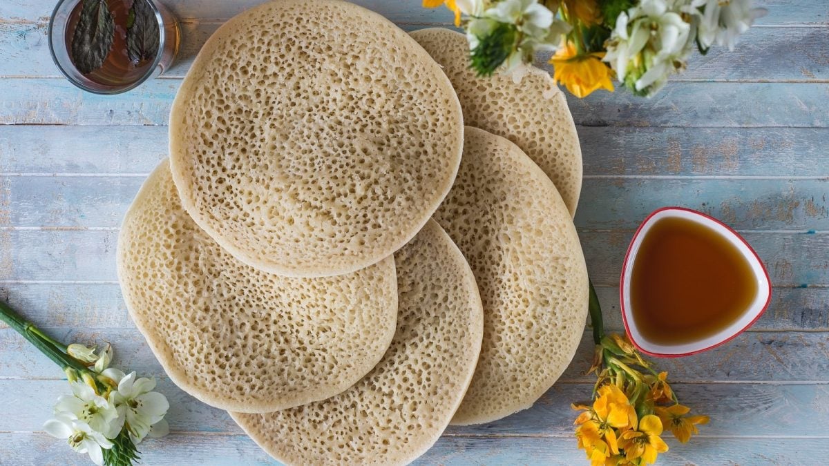 Fünf Baghrir, marokkanische Pfannkuchen aus Grieß mit Luftbläschen auf der Oberseite und fluffigem Teig, liegen gefächert aufeinander, neben einer Schüssel Honigsirup und einem Glas Minz-Tee, von oben vor hellblauem Hintergrund.