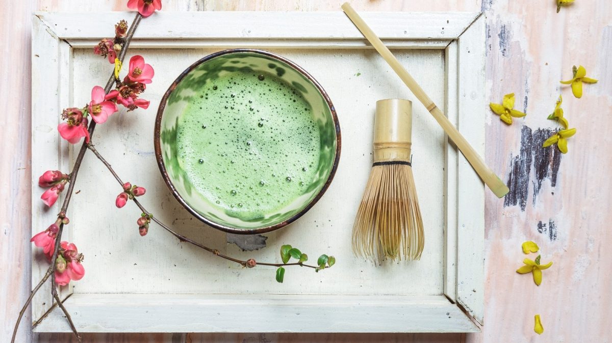 Matcha-Tee mit traditionellen Utensilien zubereitet: Der aufgeschäumte Matcha serviert in einem Chawan (Tee-Schüssel), neben einem Chasen (Tee-Besen aus Bambus) und einem Chashaku (Bambuslöffel) vor einem weißen Hintergrund mit Blüten dekoriert, von oben.