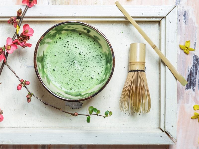 Matcha-Tee mit traditionellen Utensilien zubereitet: Der aufgeschäumte Matcha serviert in einem Chawan (Tee-Schüssel), neben einem Chasen (Tee-Besen aus Bambus) und einem Chashaku (Bambuslöffel) vor einem weißen Hintergrund mit Blüten dekoriert, von oben.