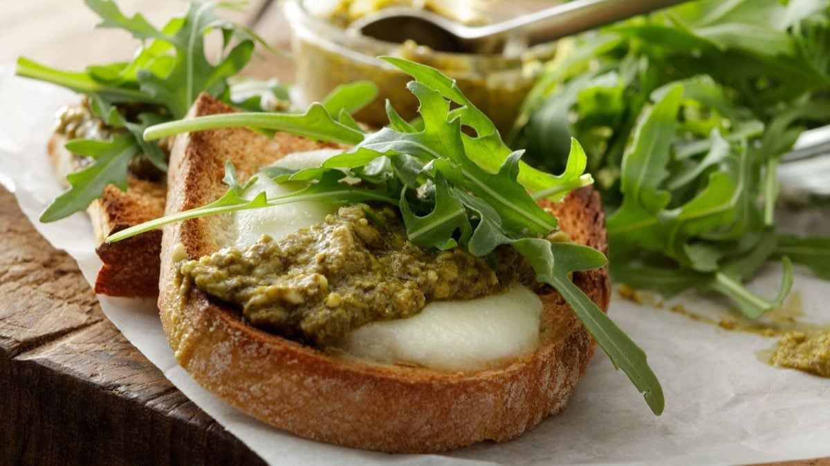 Zwei mit Ziegenkäse überbackene Crostini mit grünem Pesto und Rucola auf einem mit Papier ausgelegten Holztisch von der Seite.