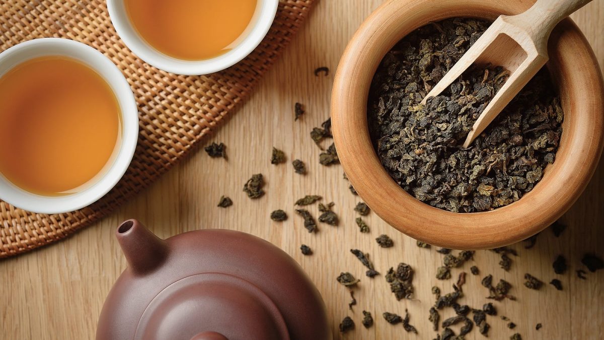 Eine Holzschale mit losen Oolong Tee Blättern neben einer Ton-Teekanne und zwei Porzellan Teetassen gefüllt mit dem gold-gelben Tee, von oben.