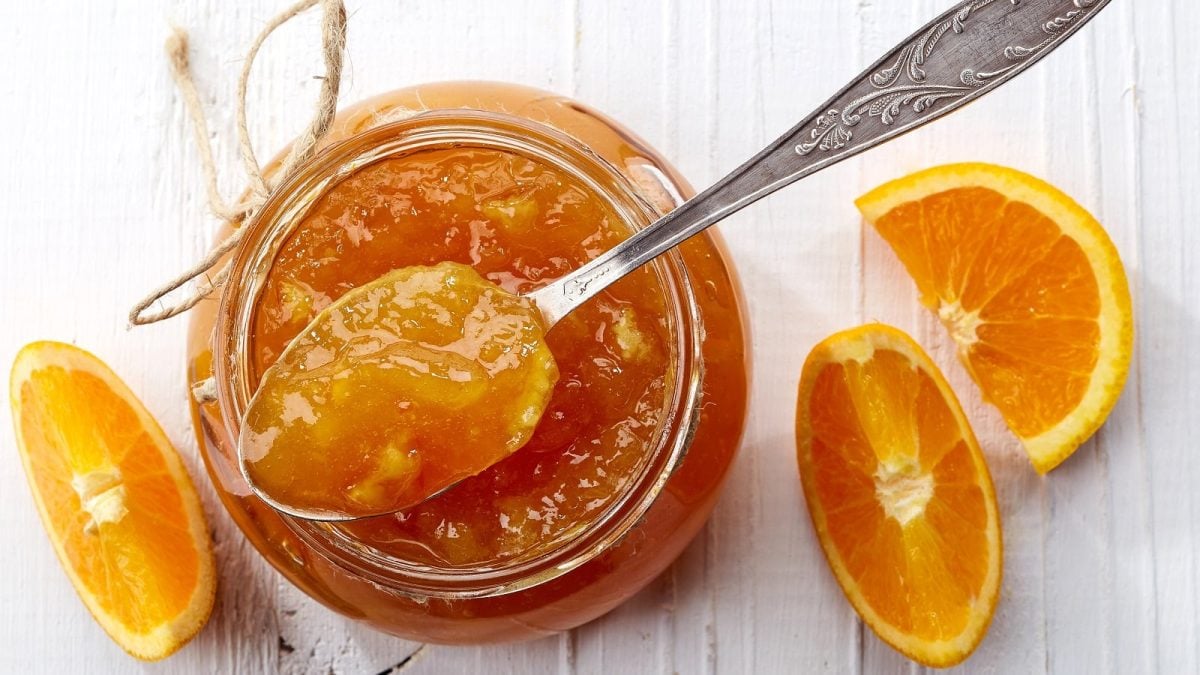 Ein offenes Glas Orangen-Grapefruit-Marmelade mit Schuss. Ein Teelöffel entnimmt dem Glas ein wenig der Marmelade. Orangen-Scheiben liegen zur Dekoration neben dem Glas, alles von oben fotografiert.