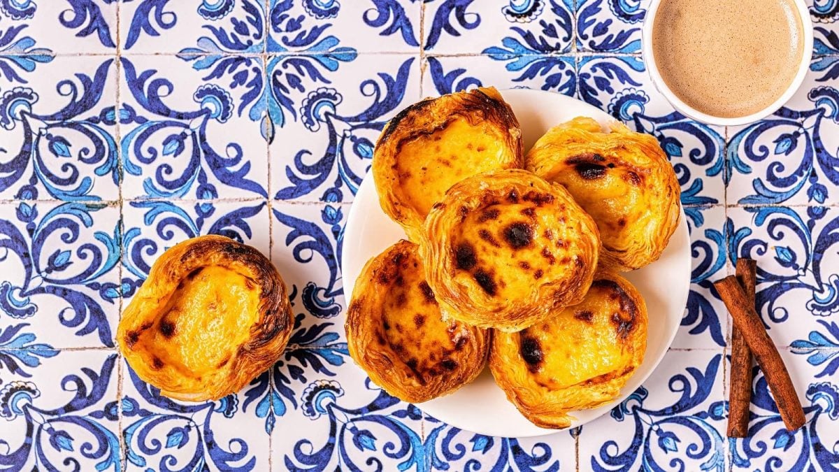Knusprig gebackene Pastéis de Nata auf einem weißen Teller gestapelt neben einer Tasse Kaffee und zwei Zimtstangen, von oben, vor einem Hintergrund mit portugiesischen Fliesen.