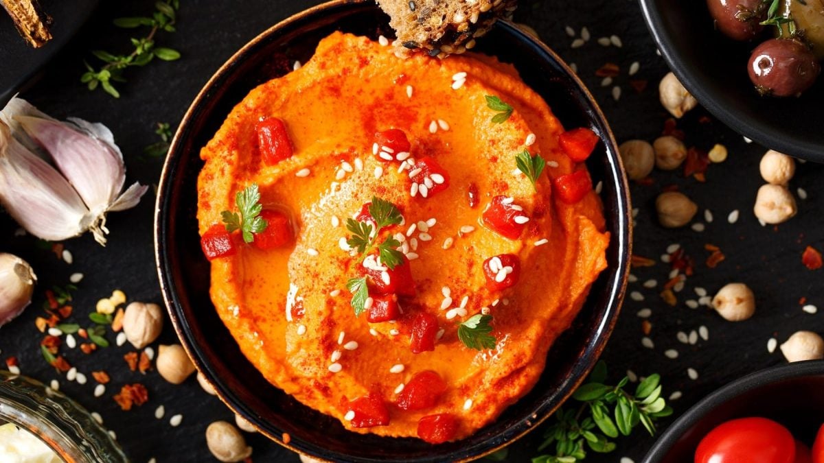 Pikanter Paprika-Hummus mit Sesam in einer dunklen Schale. Daneben einige Gewürze auf einem dunklen Untergrund.