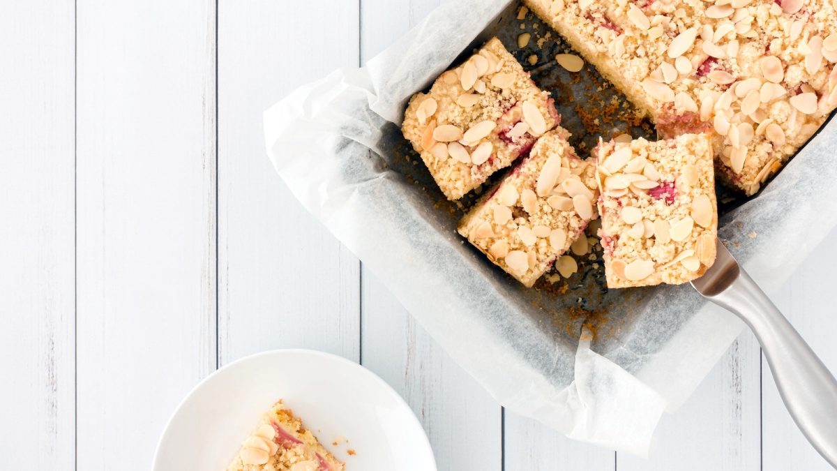 Eine eckige Backform mit einem Rhabarber-Streuselkuchen mit Mandeln, teilweise in Stücke geschnitten und ein Teller mit einem Stück Kuchen auf hellem Untergrund von oben.