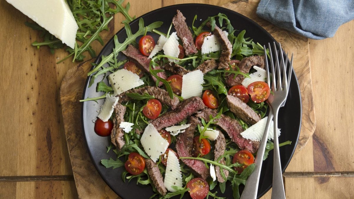 Rumpsteak mit Tomaten-Rucolasalat mit Parmesan in einem tiefen Teller, daneben zwei Gabeln, ein Stück Parmesan und ein Geschirrtuch
