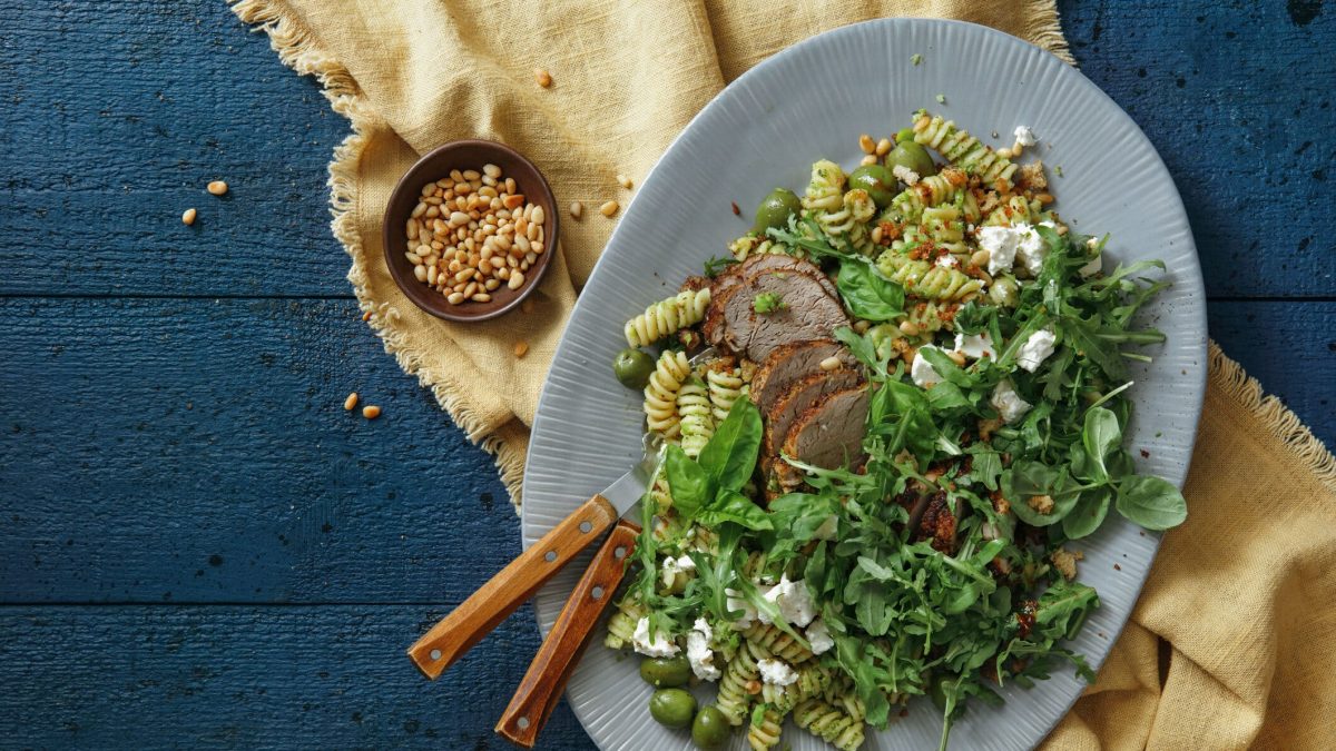 Schweinefilets mit griechischem Nudelsalat und Basilikum auf einem blauen Teller, darunter ein Tuch und daneben eine Schale mit Pinienkernen. Alles auf dunklem Untergrund.
