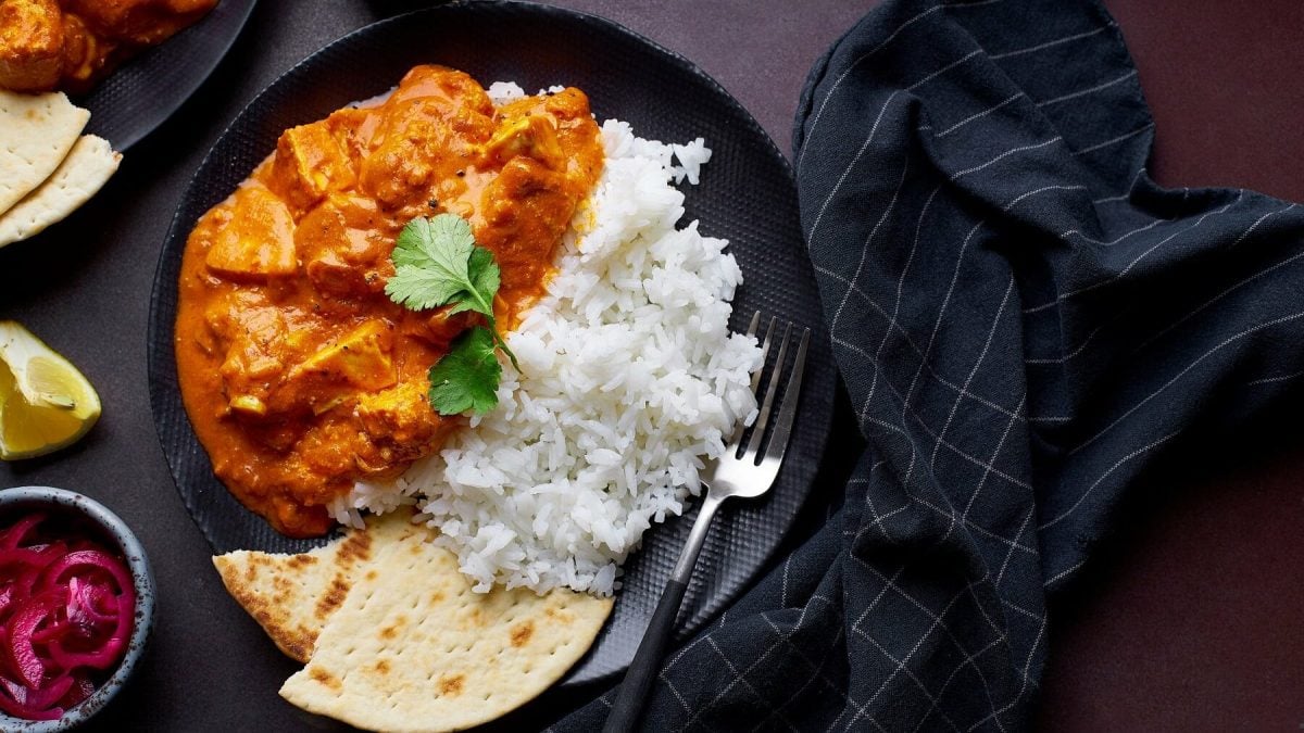 Tofu Tikka Masala mit Reis und Brot auf einem schwarzen Teller mit einer Gabel auf einem dunklen Tisch neben einem schwarzen Tuch mit streifen.