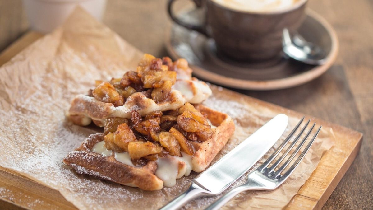 Zwei Vanille-Waffeln mit Bratapfel liegen auf einem braunen Papier auf einem Holzbrett. Neben den Waffeln liegen Messer und Gabel. Im Hintergrund steht ein weißer Becher und eine braune Tasse mit Kaffee. Die Tasse steht auf einer passenden, braunen Untertasse.