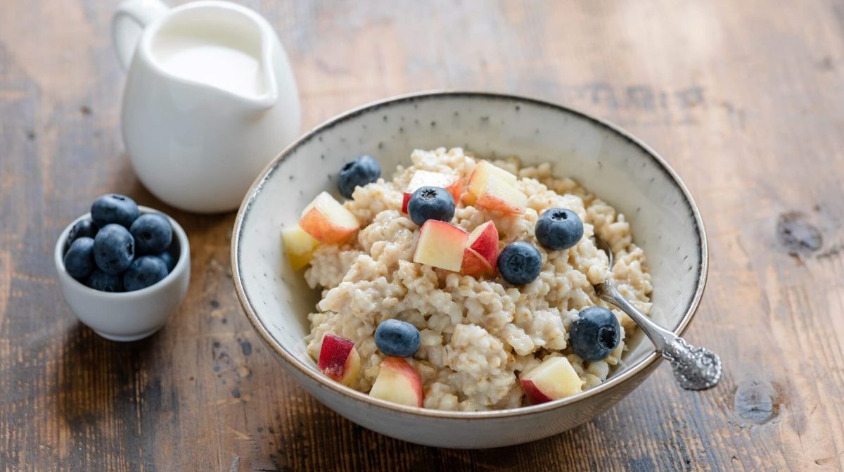 Veganes Bircher Müsli mit Blaubeeren und Pfirsich in einer hellen Schüssel auf einem Holztisch neben einem Kännchen mit Milch und einer kleinen Schüssel mit Blaubeeren.