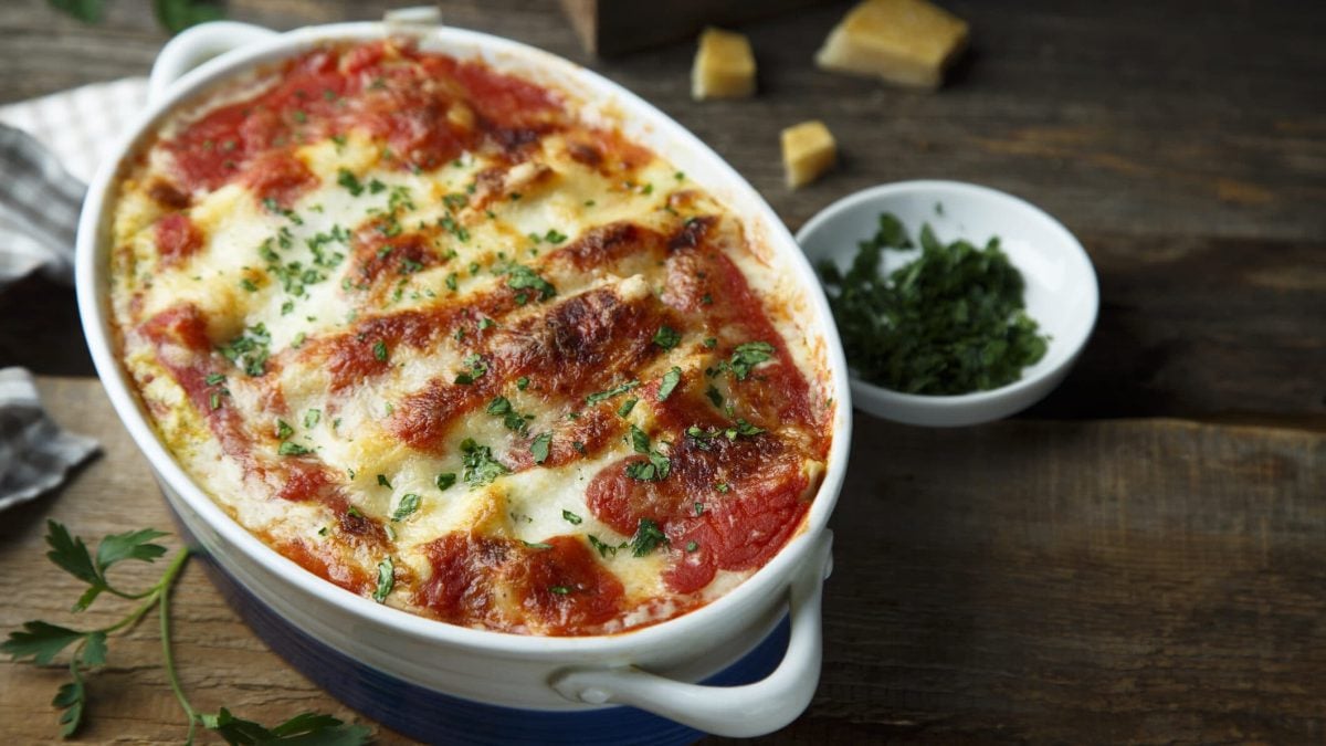 Vegetarische Cannelloni überbacken in einer Auflaufform, daneben eine Schale mit Kräutern und ein paar Stücke Parmesan.