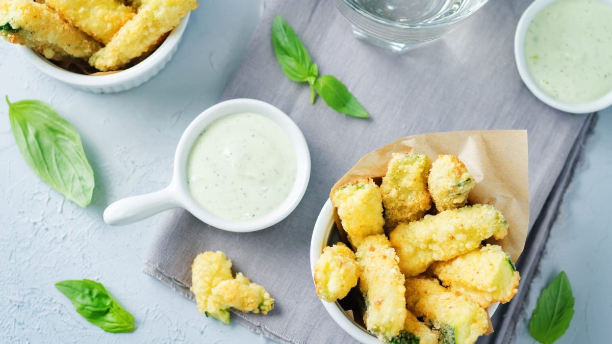 Hellblauer Untergrund mit einer Schale Zucchini-Pommes umgeben von Dips.