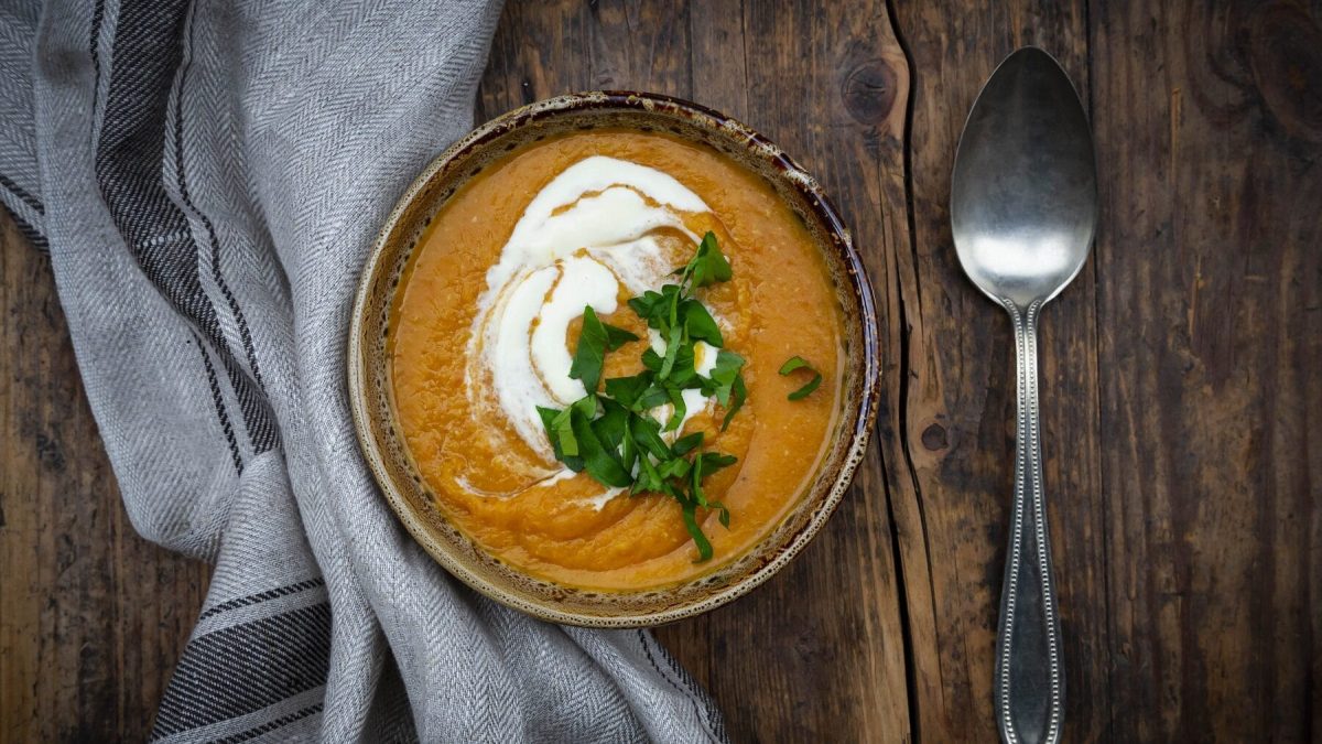 Rote Linsensuppe mit Créme Fraiche und frischer Petersilie in einer gemusterten Schüssel auf einem dunklen Holztisch neben einem grauen Tuch un einem Löffel.