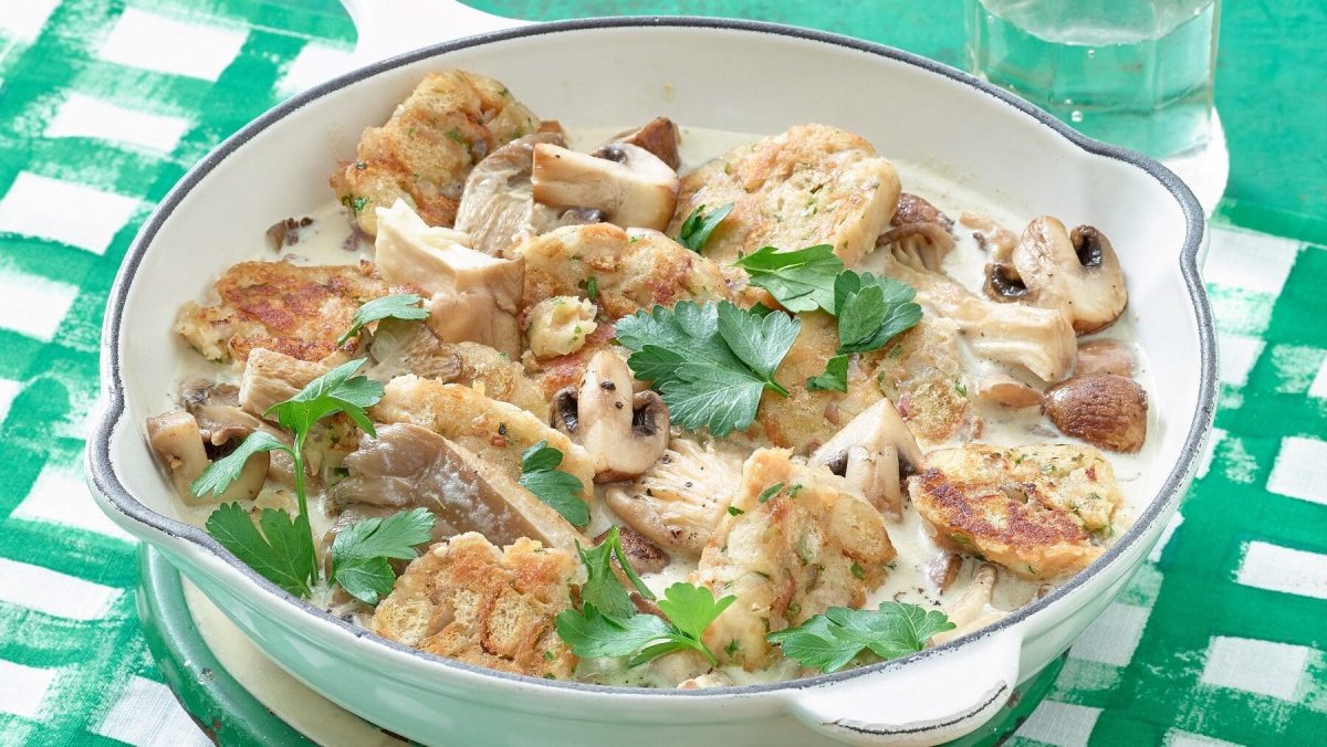 Eine weiße Pfanne mit veganem Semmelknödel-Geschnetzelten mit Pilzen steht auf einem weiß-grünen Untergrund. Rechts oben neben der Pfanne steht ein Glas mit Wasser. Das Geschnetzelte ist garniert mit Petersilie.