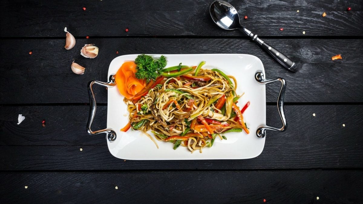 Eine Portion asiatischer Bratnudeln liegt auf einem weißen Tablett mit silbernen Griffen auf einem schwarzen Holztisch. Neben dem Tablett liegt ein Löffel und drei Knoblauchzehen. Auf dem Tisch sind Pfefferkörner verteilt.