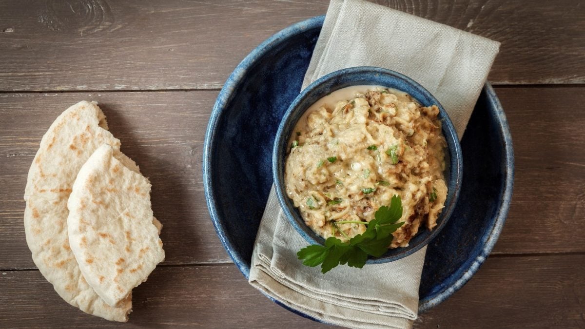 Eine blaue Schüssel mit Baba Ghanoush gefüllt und Petersilie verziert neben zwei angebrochenen Stücken Fladenbrot, von Oben vor einem Holzhintergrund.
