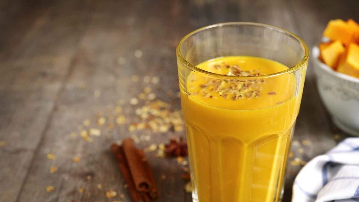 Bananen-Kürbis-Smoothie im Glas auf einem Holztisch, daneben Zimtstangen und Sternanis, ein Geschirrtuch und eine Schale mit Kürbisstücken.
