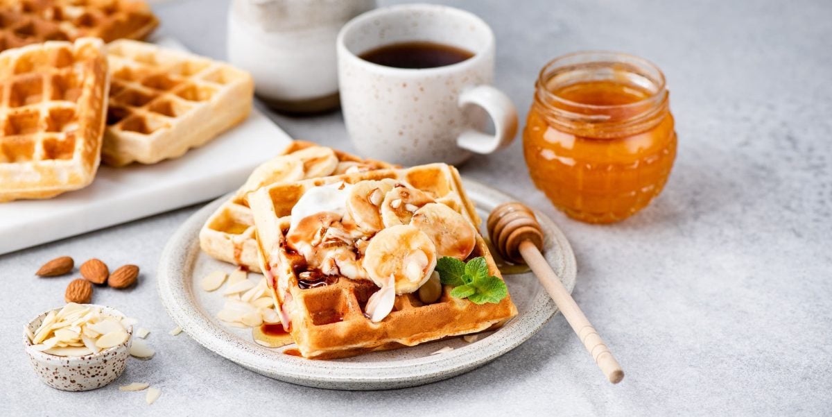 Bananewaffeln mit Bananenscheiben, Mandeln, Joghurt und Minze auf einem grauen Teller neben einem Honiglas, einer Kaffeetasse und einem Brett mit Waffeln auf einem grauen Steintisch.