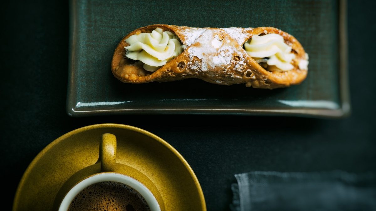 Ein sizilianisches Cannoli mit Puderzucker bestäubt auf einem rechteckigen, dunklen Teller neben einer Tasse Espresso vor dunklem Hintergrund von oben.
