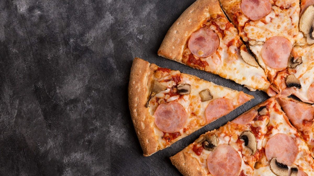 Eine Champinon-Pizza mit Speck liegt auf einem dunklen Untergrund aus Stein. Die Pizza ist bereits angeschnitten und ein Stück steht ein Wenig hervor.