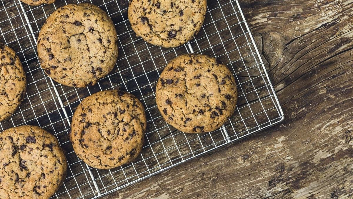 Mehrere runde Chocolate-Chip-Cookies liegen auf einem silbernen Gitter auf einem braunen Holztisch.