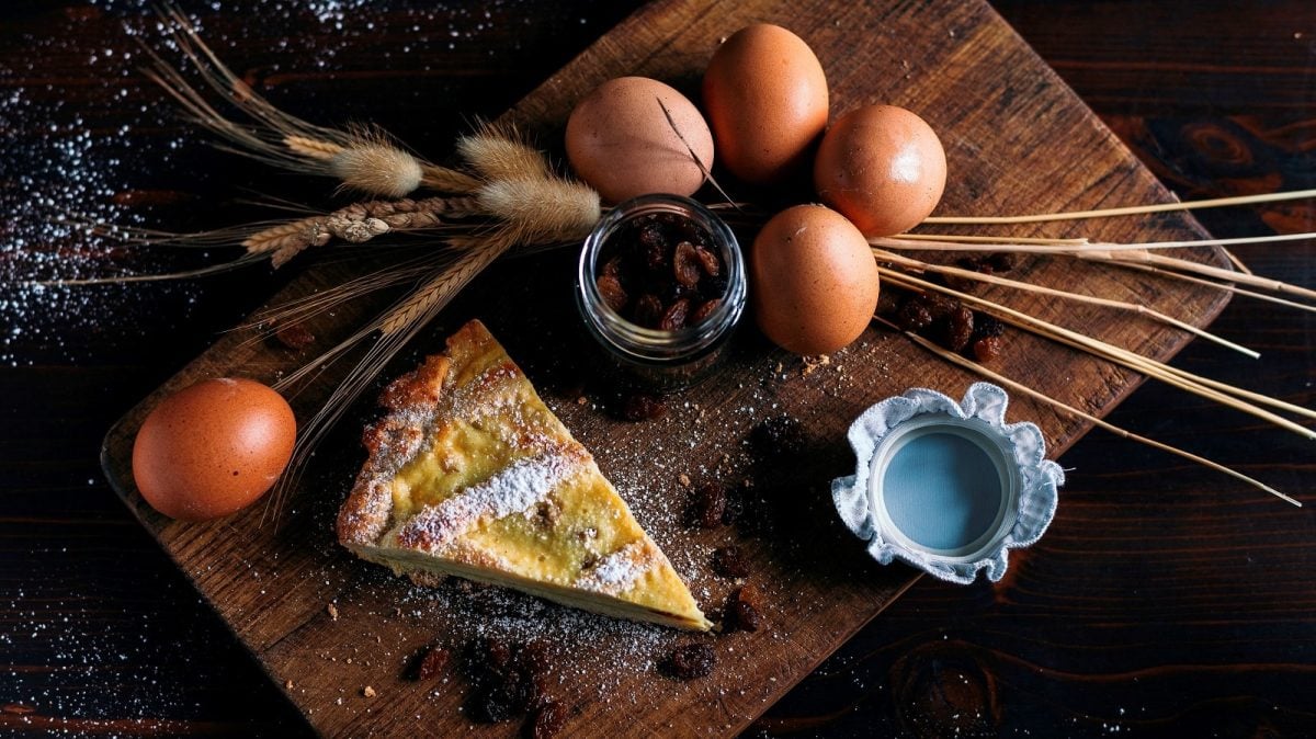 Ein Stück der Crostata Ricotta e Visciole mit Puderzucker bestreut, neben Eiern, einem Glas mit Kirschen und getrockneten Weizenstängeln, auf einem Holzbrett von Oben.