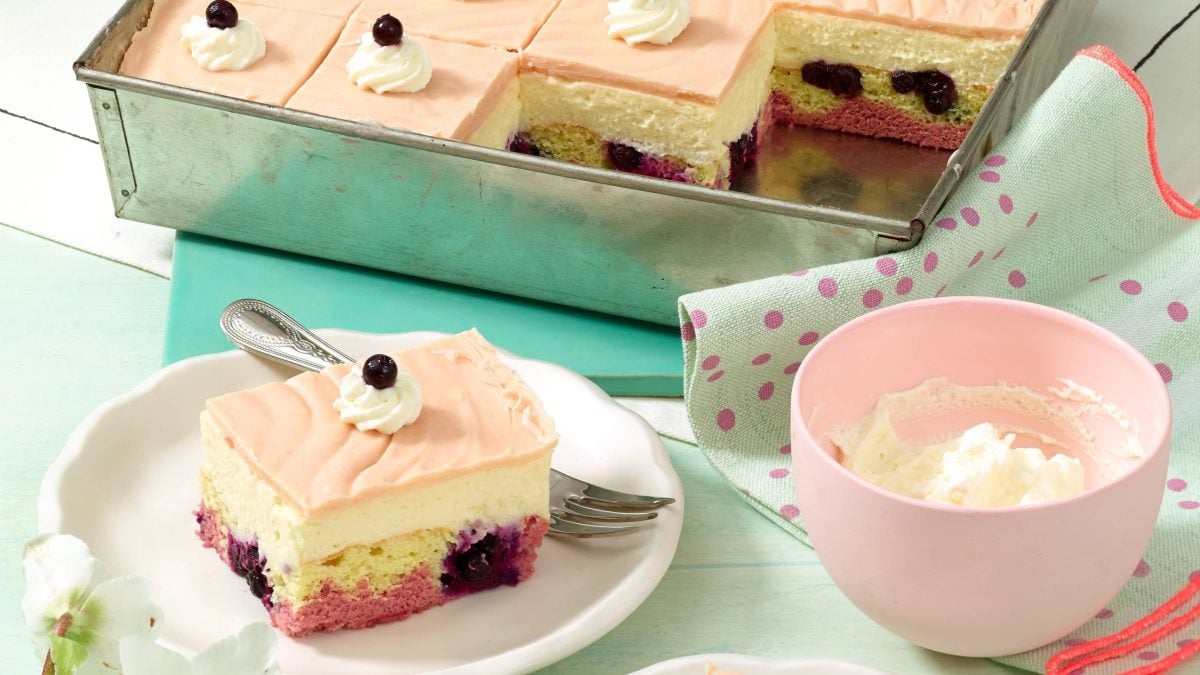 Donauwelle mit Marzipan aus einem tiefen Blech auf einem weißen Teller neben einer rosa Schüssel mit Sahne auf einer grünen Tischdecke.