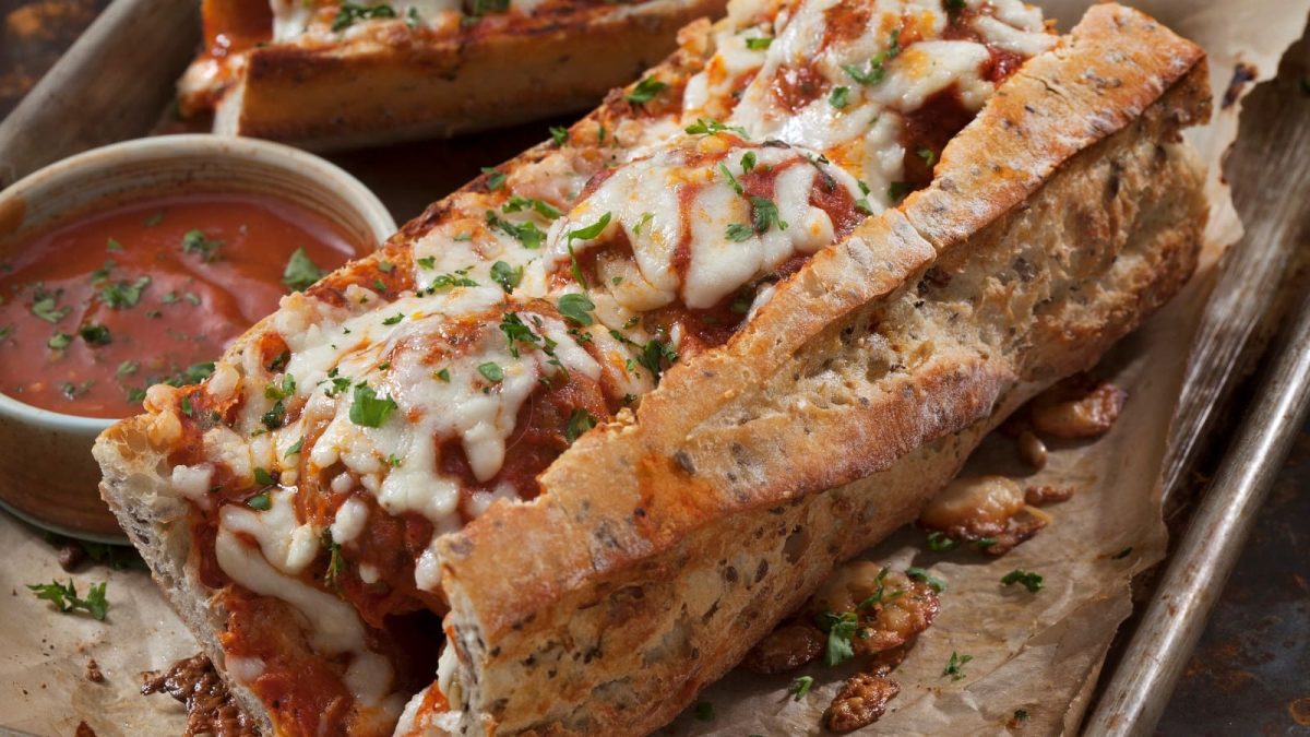 Zwei überbackene Baguette-Sandwiches mit Fleischbällchen und Tomatensauce, garniert mit Basilikum neben einer kleinen Schüssel Tomatensoße.